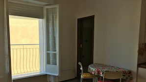 Una habitación pequeña con balcón, una mesa con mantel floral, dos sillas y una puerta.