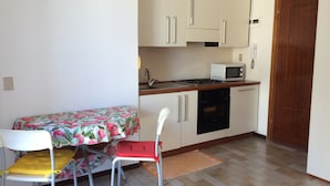 Cocina pequeña con microondas, estufa y lavamanos. Se ve una mesa con sillas y una puerta a la derecha.