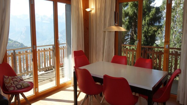 A dining area with a large table and red chairs, large windows with a view of mountains, and a wooden balcony. - Cozy Apartment for Groups (Sion)