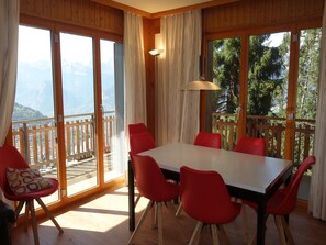 A dining area with a large table and red chairs, large windows with a view of mountains, and a wooden balcony. - Cozy Apartment for Groups (Sion)