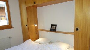 A single bed with white bedding, a wooden headboard, a shelf with a framed picture, and a window with a wooden frame. - Cozy Apartment for Groups (Sion)