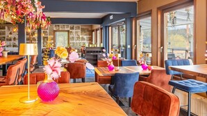 A modern restaurant interior with wooden tables, cushioned chairs, and large windows offering views of the outdoors. - Achouffe Valley (Houffalize)