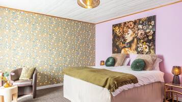 Une chambre à coucher avec un papier peint à motifs floraux, un grand lit avec de nombreux coussins, une chaise, une table d’appoint et un tableau mural.