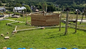 A wooden play structure in a grassy field with a view of houses and hills in the background. - Achouffe Valley (Houffalize)