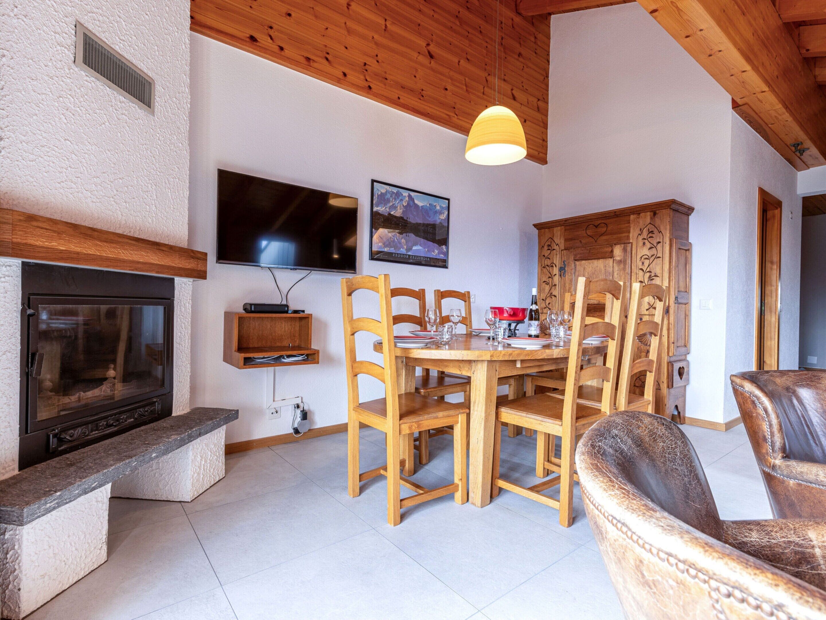 A dining area with a wooden table and chairs, a television mounted on the wall, and a fireplace.