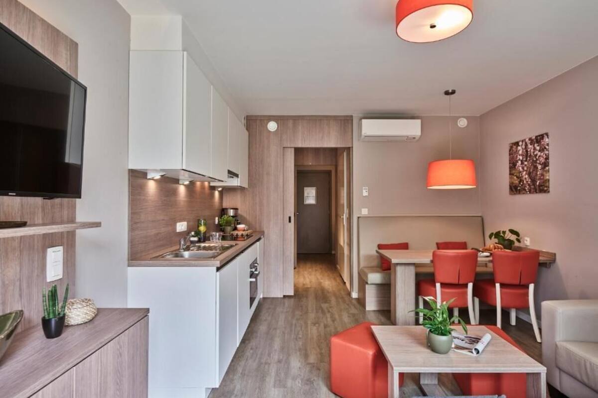 A modern apartment with a kitchen, dining area, and living room. It features a flat-screen TV, a dining table with red chairs, and a sofa.