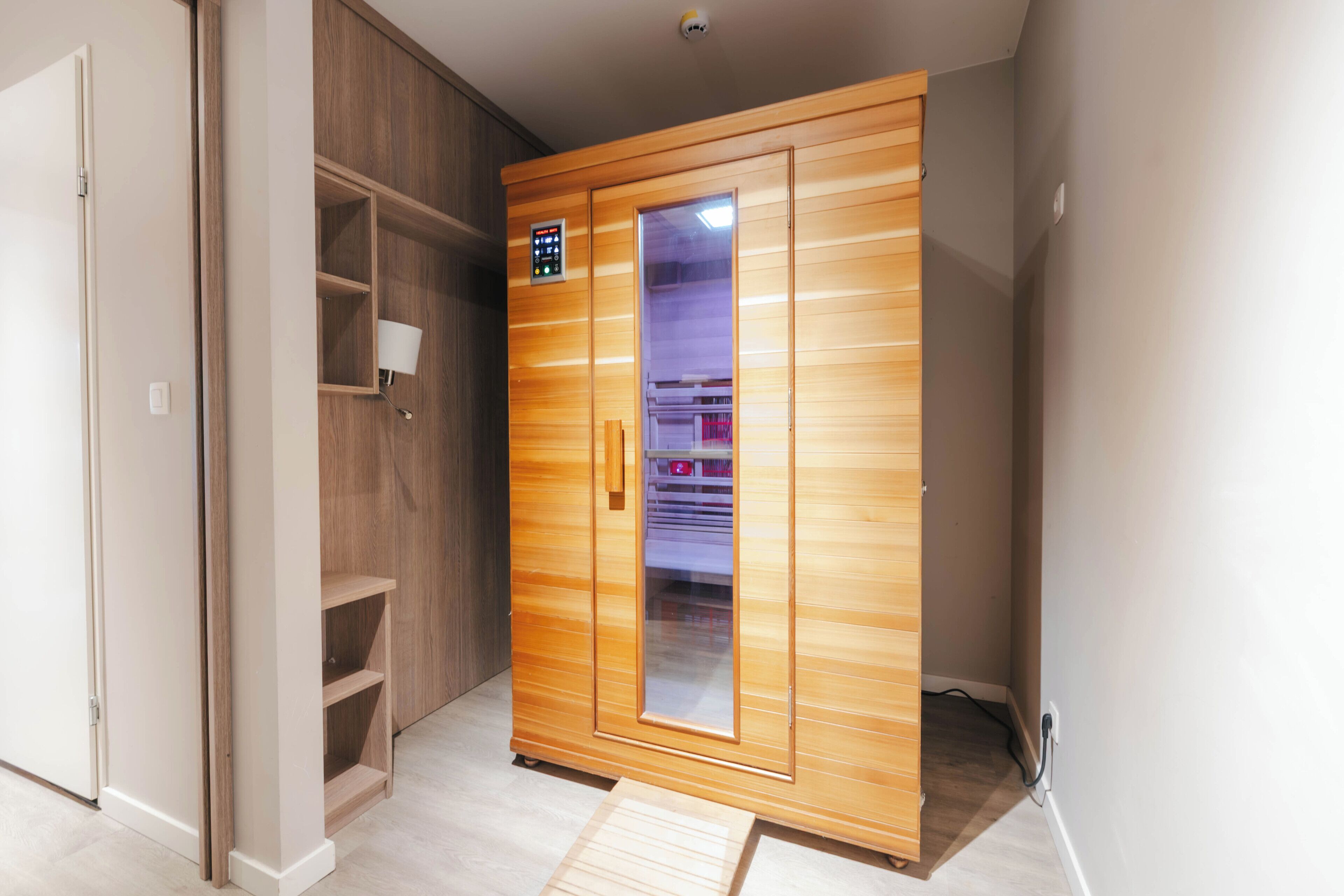 A wooden sauna with a control panel, a glass door, and a shelf with a lamp.