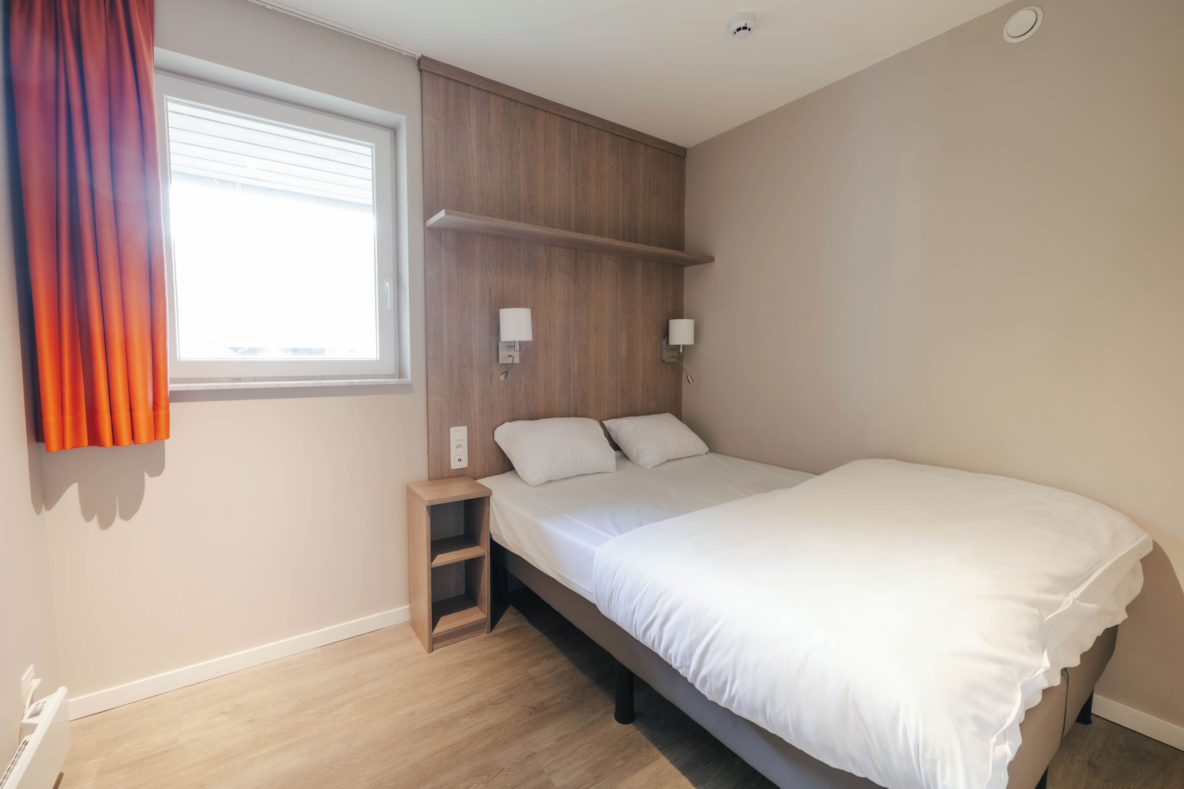 A bedroom with a bed, a nightstand, a shelf, a window with blinds, and a wall-mounted lamp.