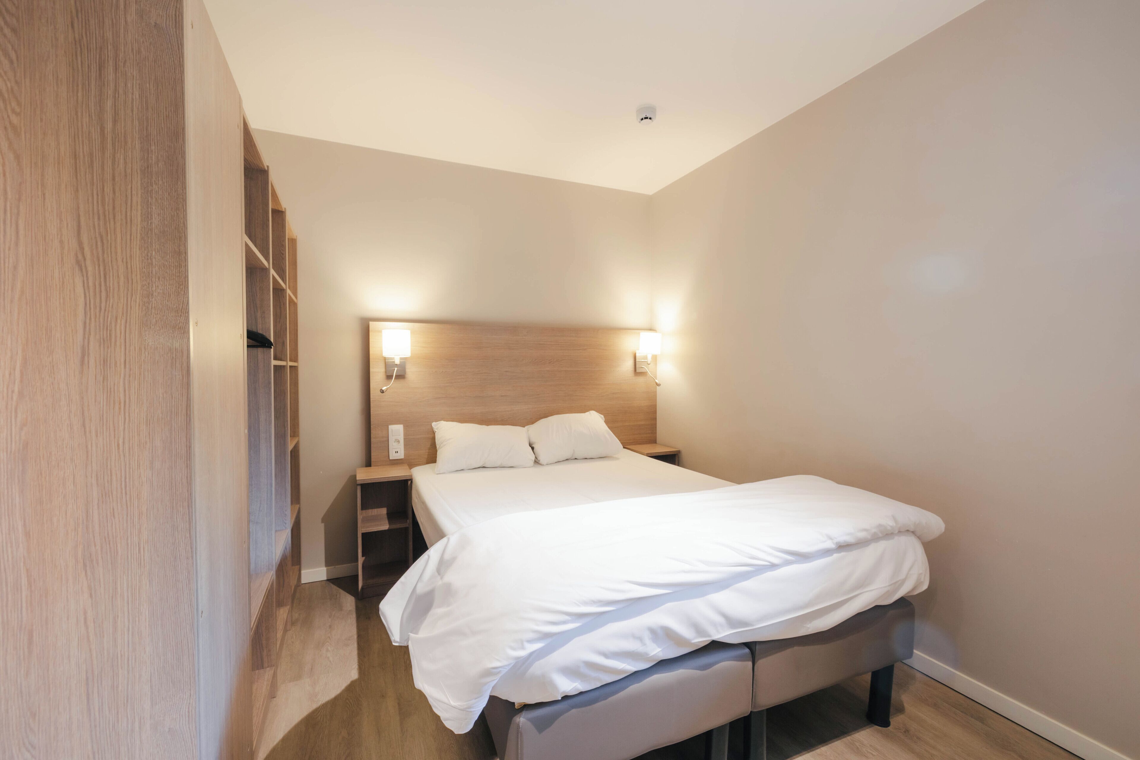 A modern hotel room with a large bed, bedside tables, and built-in shelving.