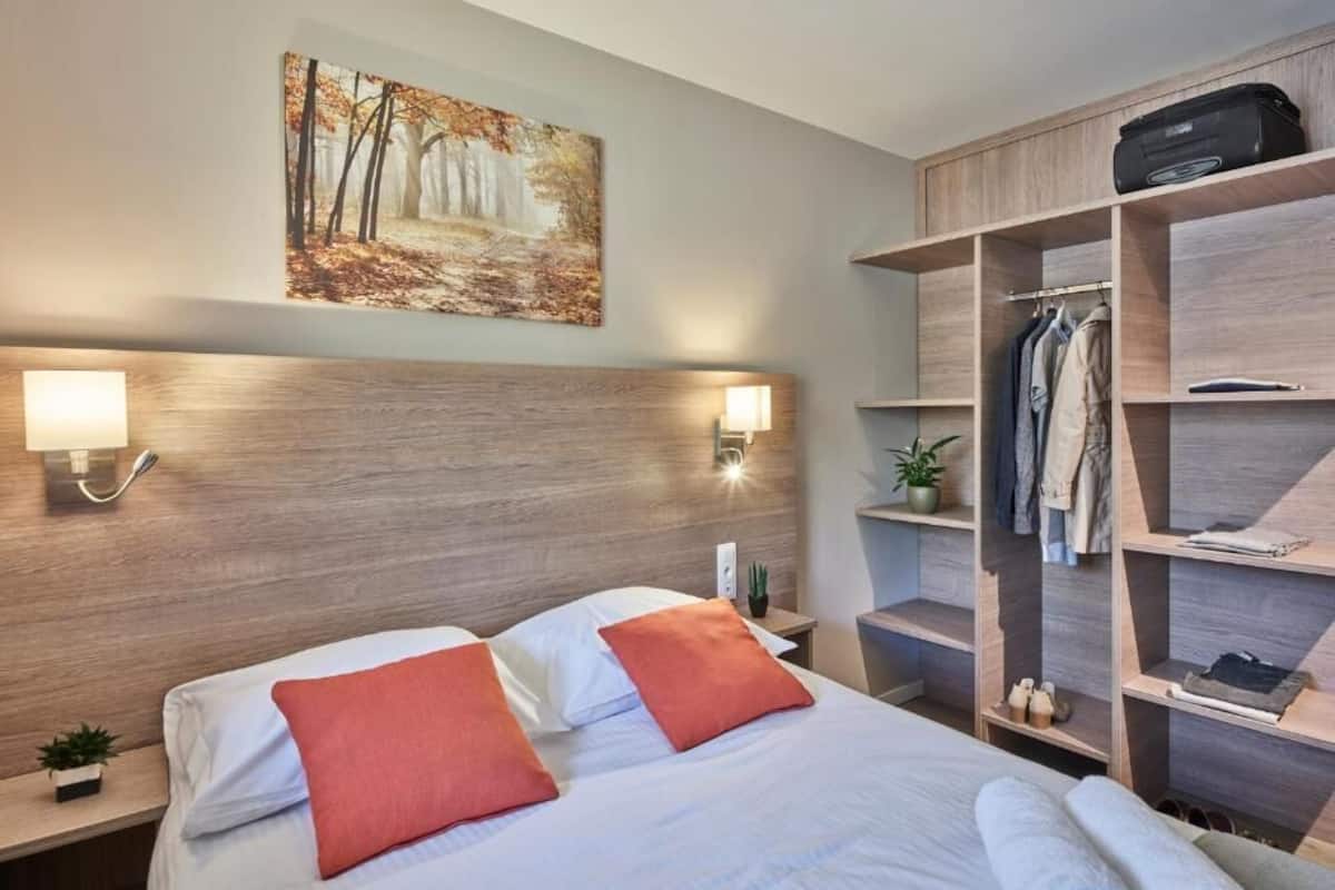 A hotel room with a wooden headboard, a bed with white linens and orange pillows, a painting of a forest, a wardrobe with shelves, and a suitcase on top.