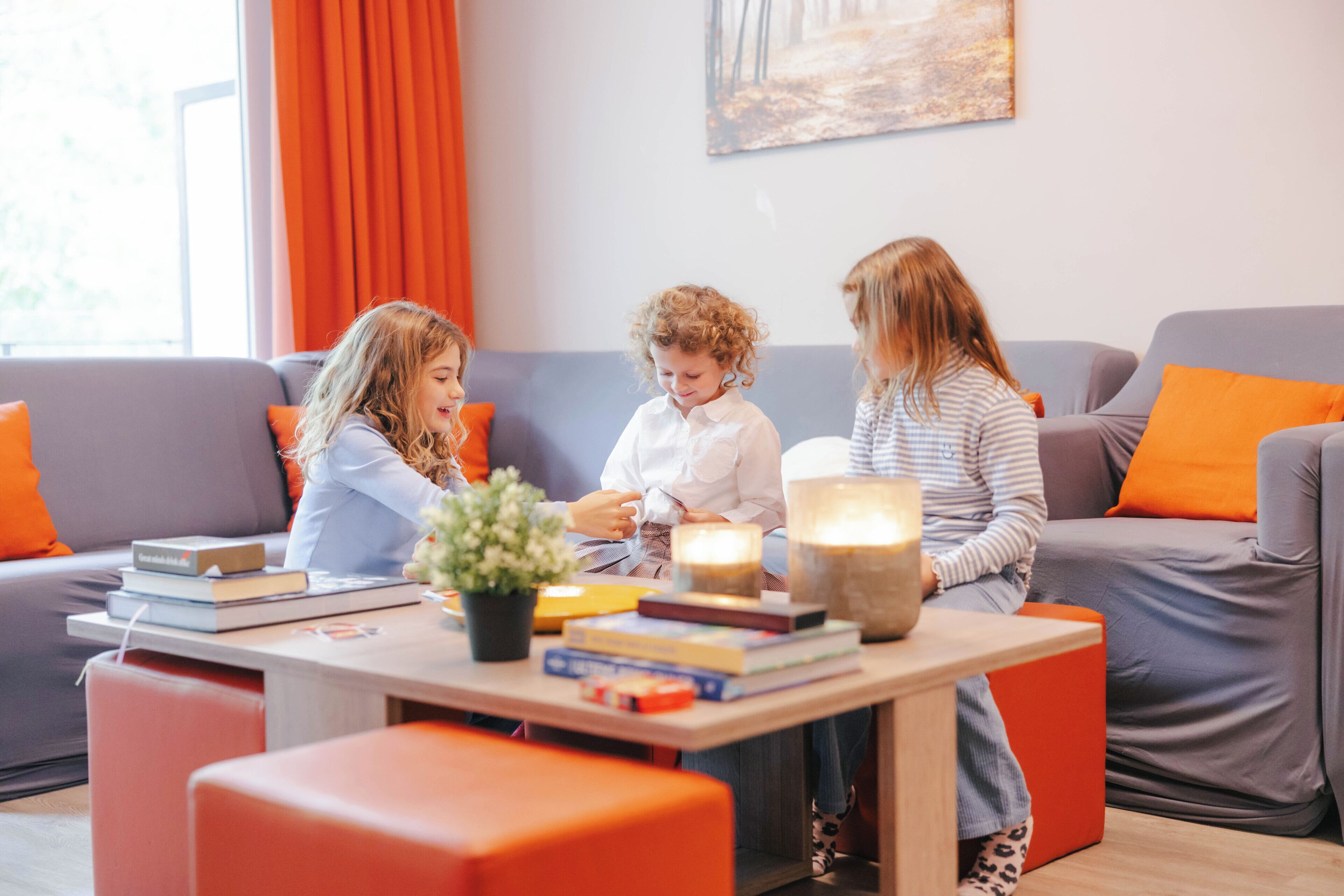 Three children sitting on a couch with books and a lamp, in a room with orange cushions and a painting on the wall.