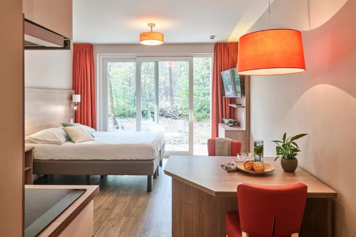 A modern hotel room with a bed, a desk with a chair, a TV, and a large window with curtains.