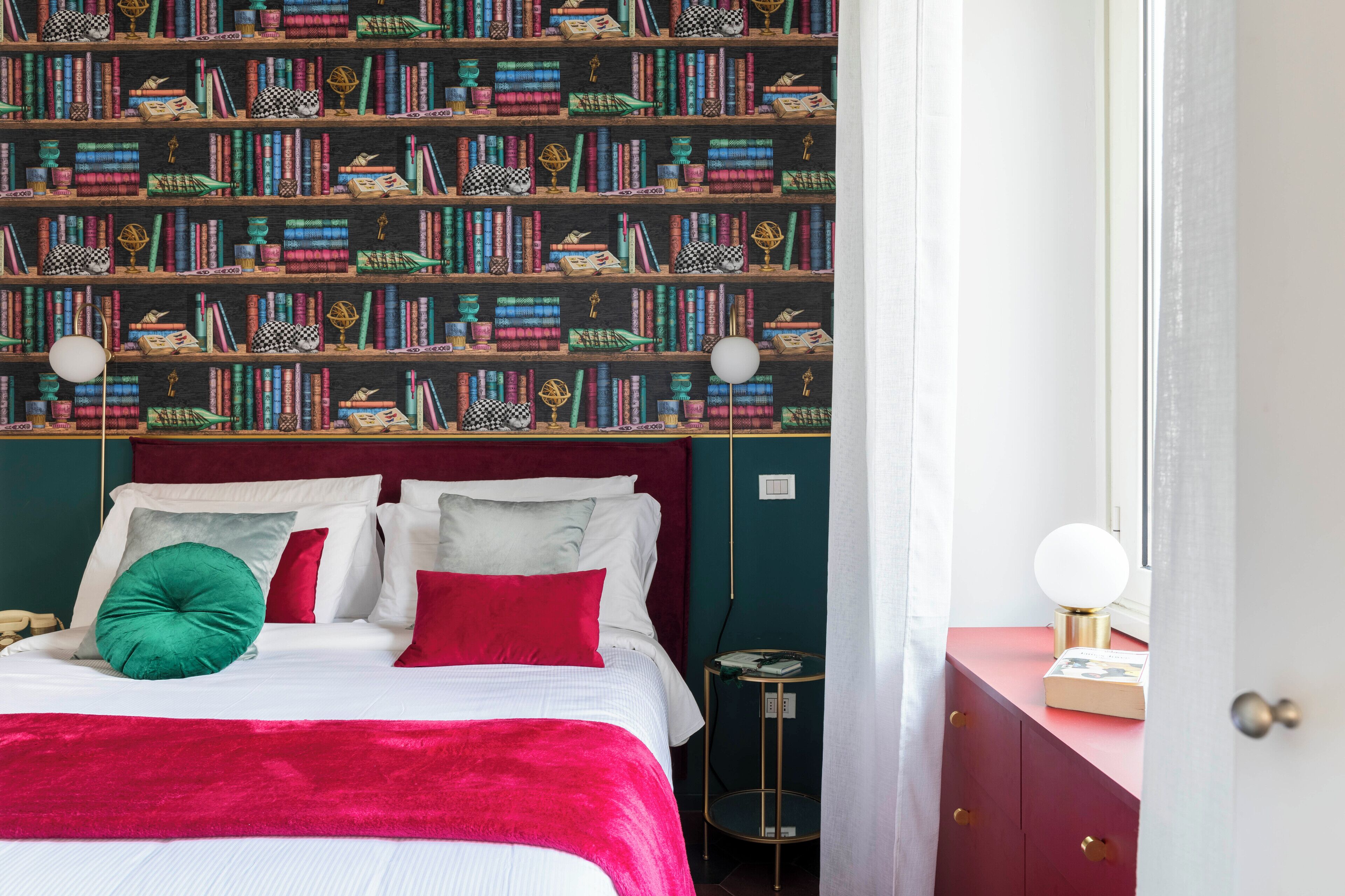 A neatly made bed with a red blanket and pillows, a bookshelf filled with books, and a small round table with a lamp.