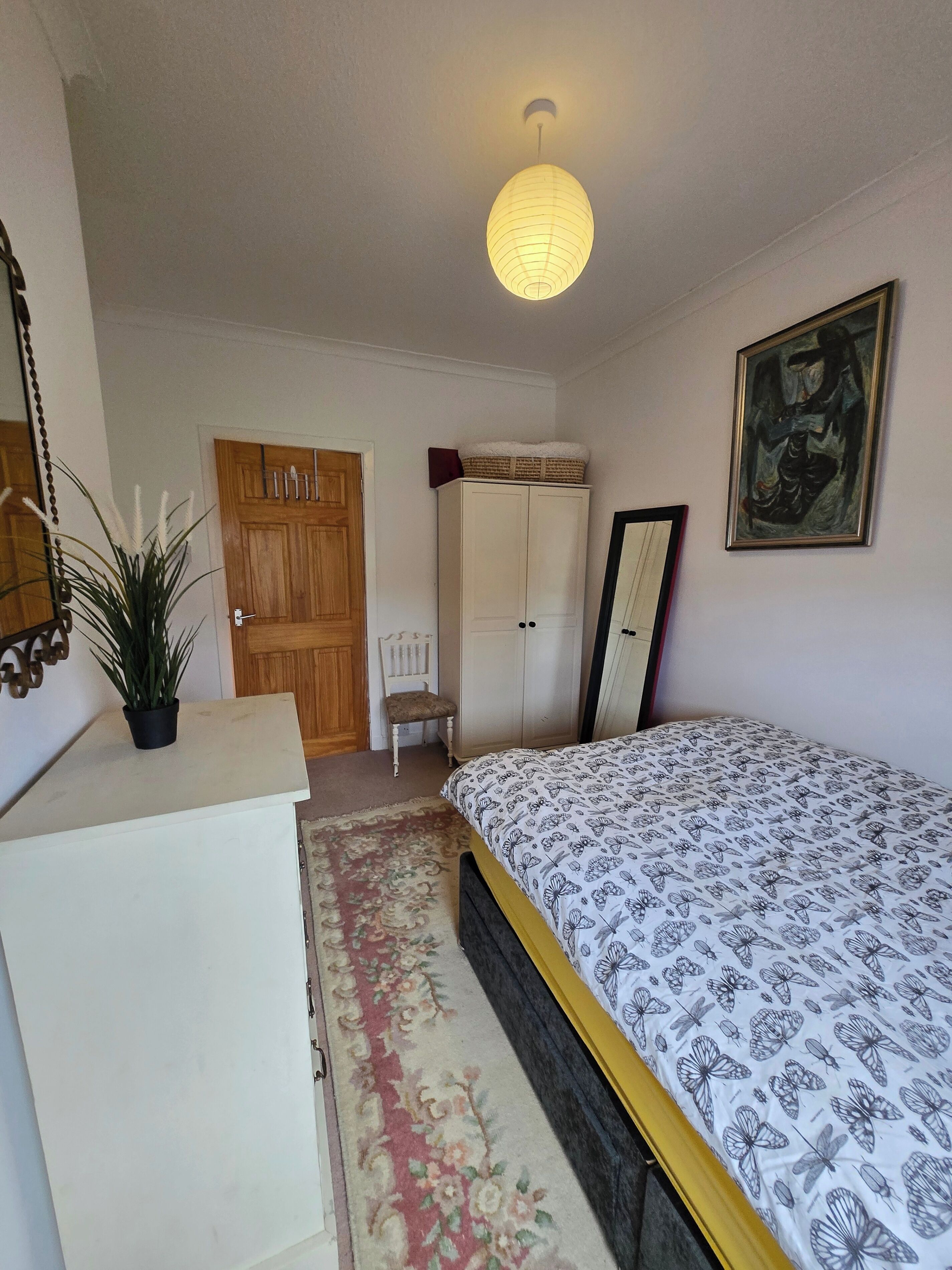A bedroom with a bed, a wardrobe, a desk, a chair, a hanging light fixture, and a painting on the wall.