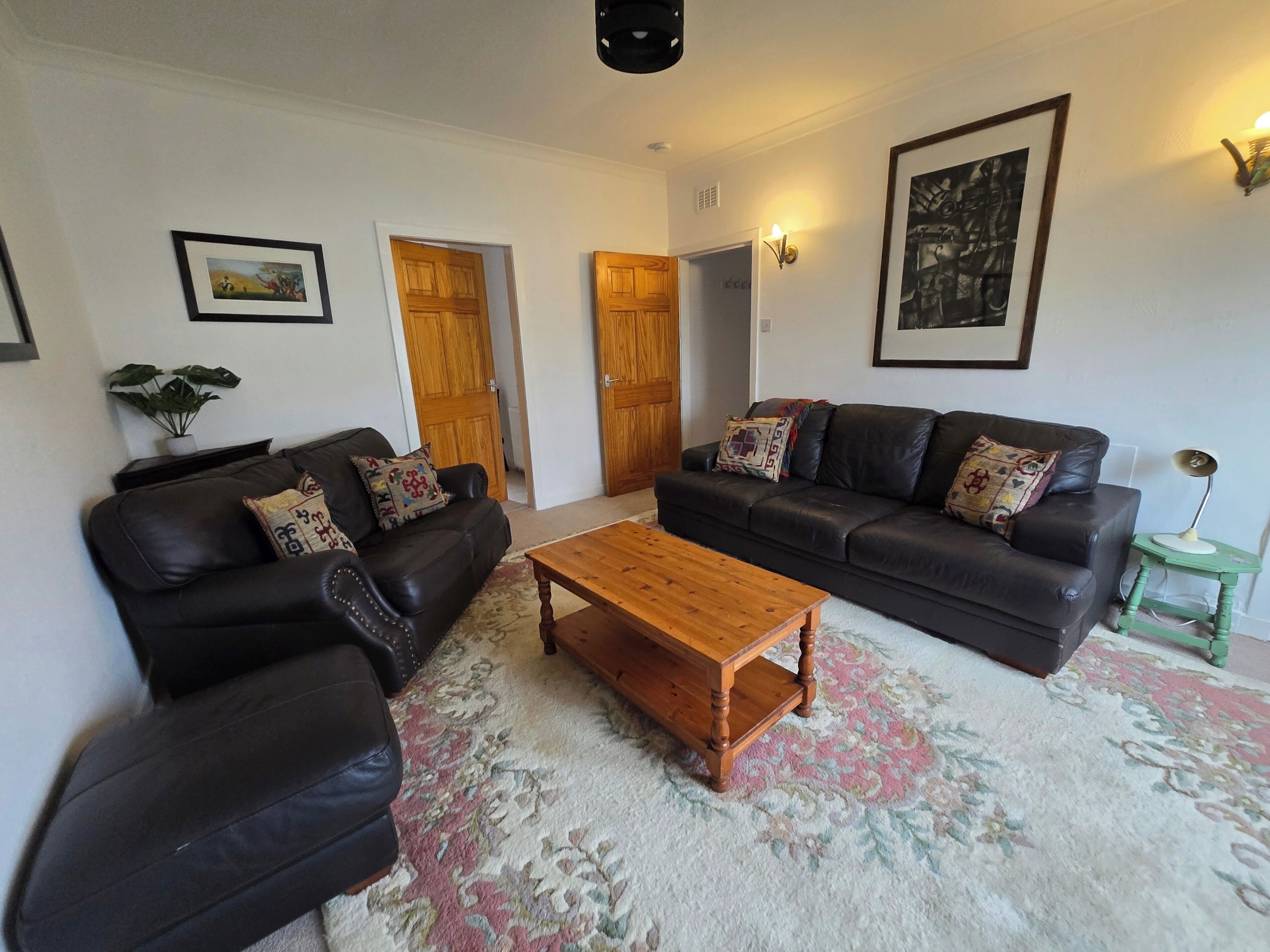 A living room with two black leather sofas, a wooden coffee table, and a patterned rug. There are doors leading to other rooms, a framed picture on the wall, and a small table with a lamp.