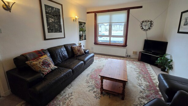 A living room with a black leather sofa, a wooden coffee table, a TV, and a large window with a view of buildings outside. - Apartmentsleeps5-parking-garden-petsok (Kelty)