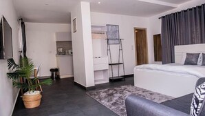 Ein modernes Hotelzimmer mit einem großen Bett, einem kleinen Schreibtisch, einem Sessel, einer Zimmerpflanze und Blick auf die Küche und den Wohnbereich.