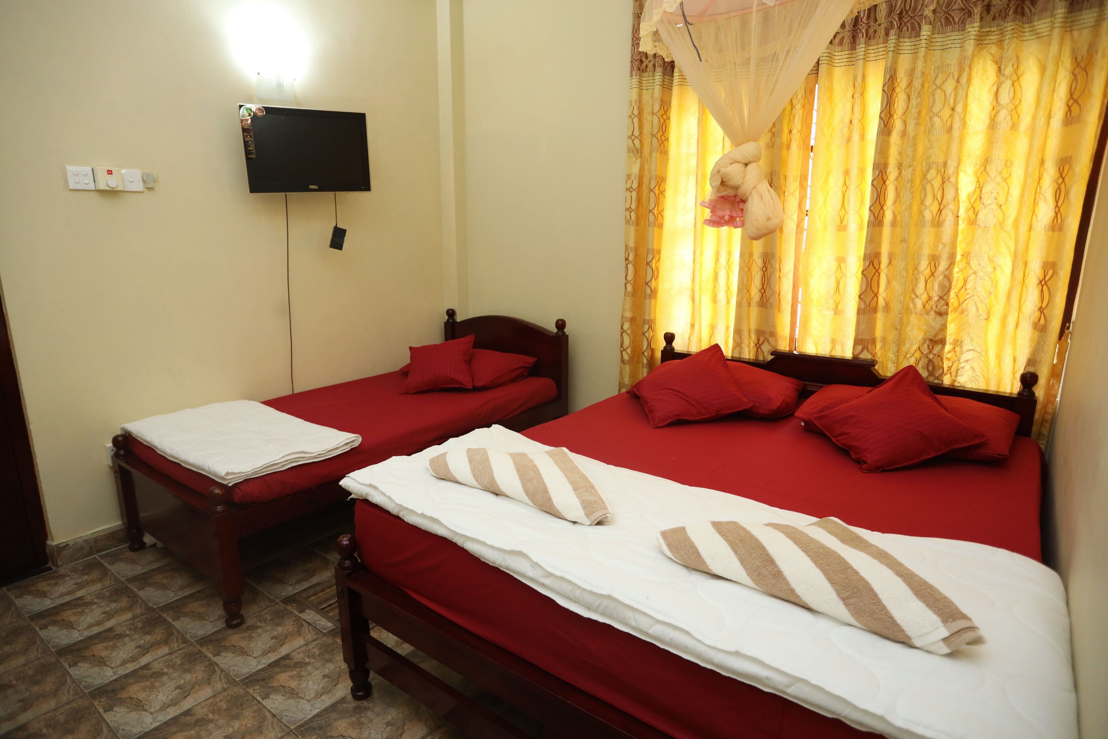 A room with two beds, each with red bedding and pillows, a flat-screen TV mounted on the wall, and a window with yellow curtains.