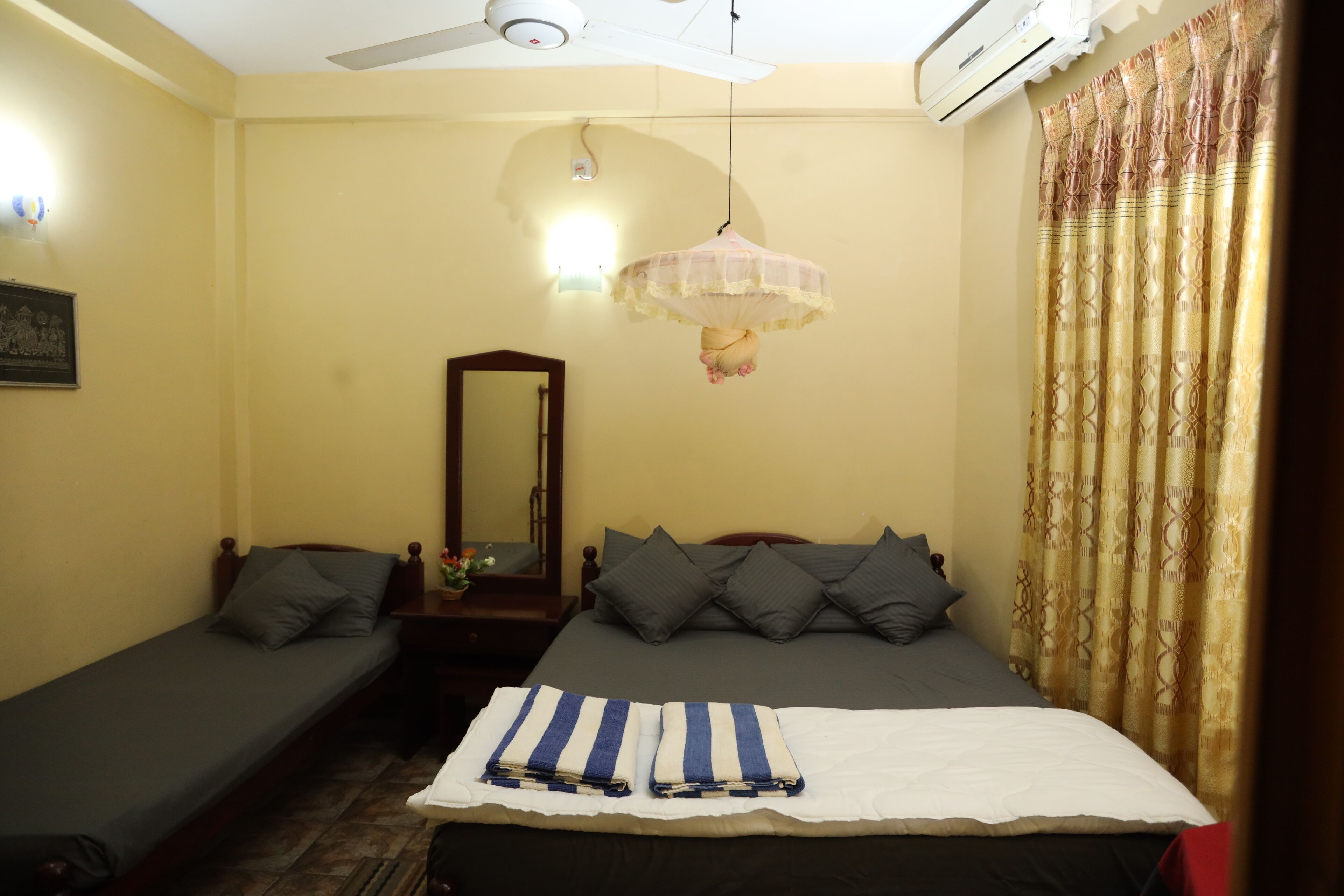 A bedroom with a single bed, a smaller bed, a ceiling fan, an air conditioning unit, a mirror, and a curtain.