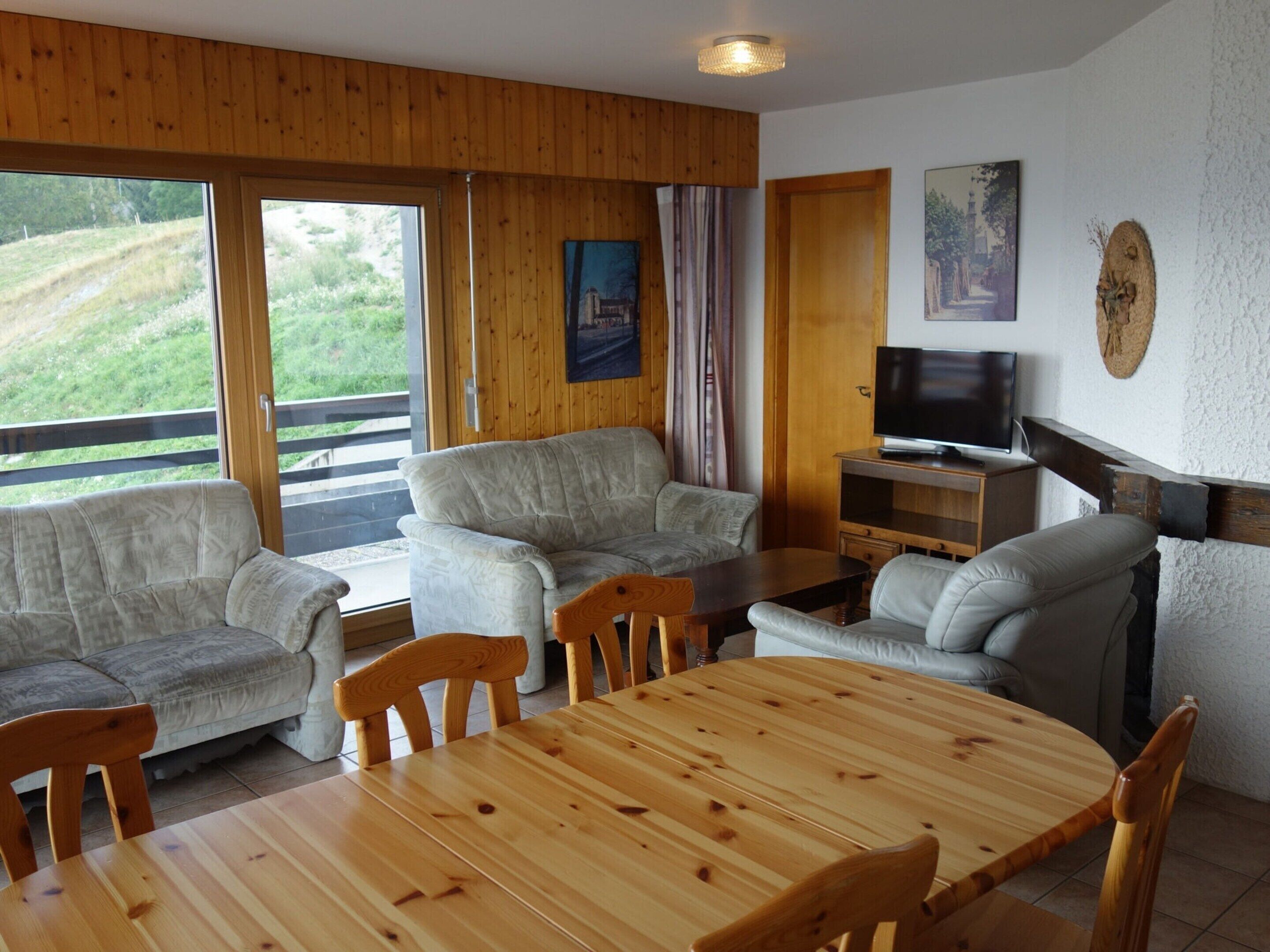 A cozy living room with a wooden table and chairs, a sofa set, a television, and a scenic view through large windows.
