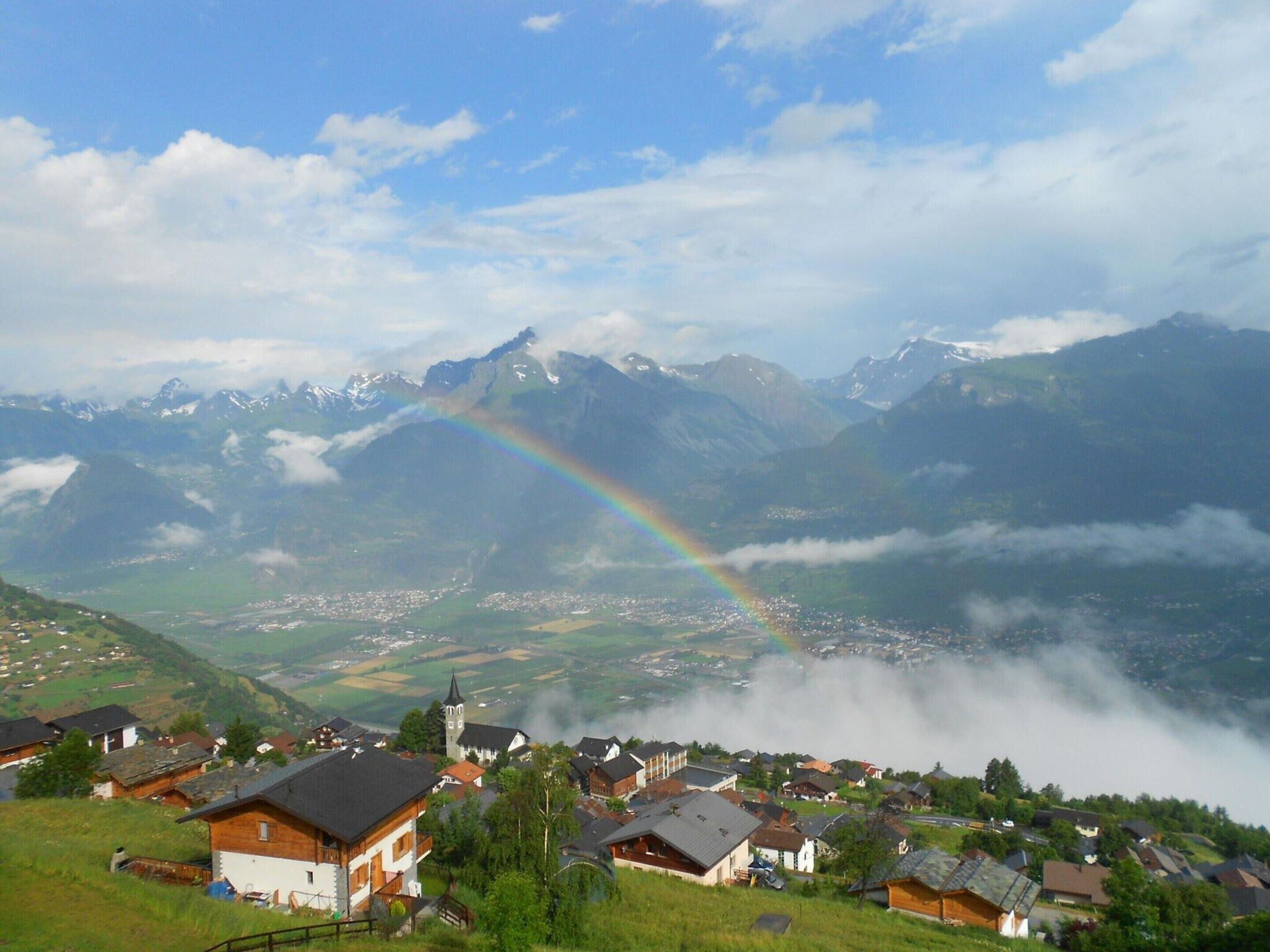 Un village de montagne aux maisons traditionnelles, avec une église et un arc-en-ciel qui surplombe la vallée.