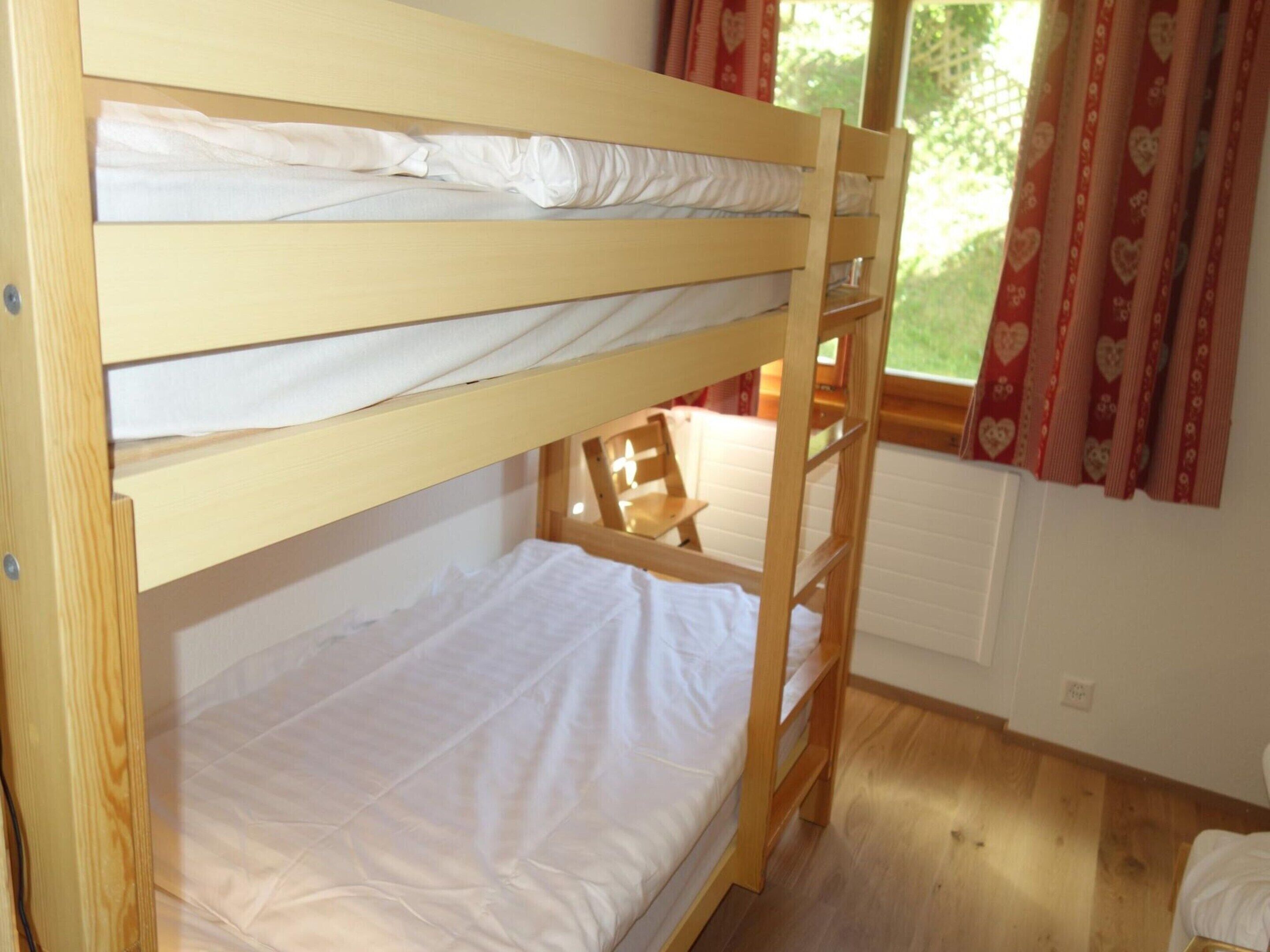 A bunk bed room with a wooden floor, a window with curtains, and a chair.
