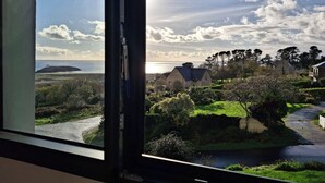 Vista costera a través de una ventana, que incluye una zona con césped, una carretera y un edificio con techo oscuro.