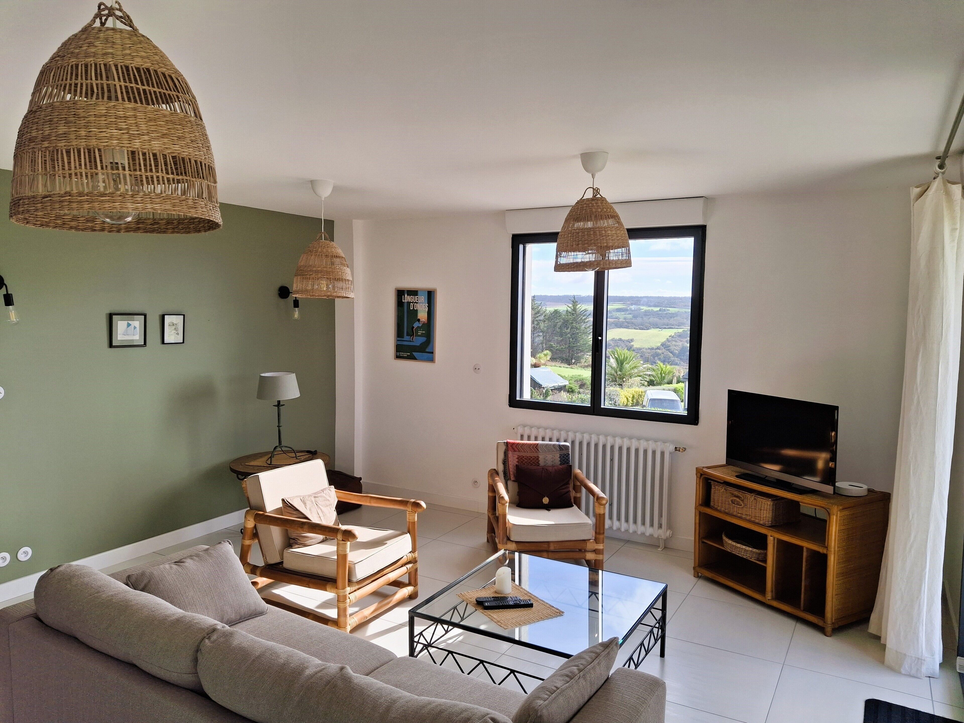A living room with a sofa, two wicker chairs, a glass coffee table, a TV, and a view of the countryside through a large window.