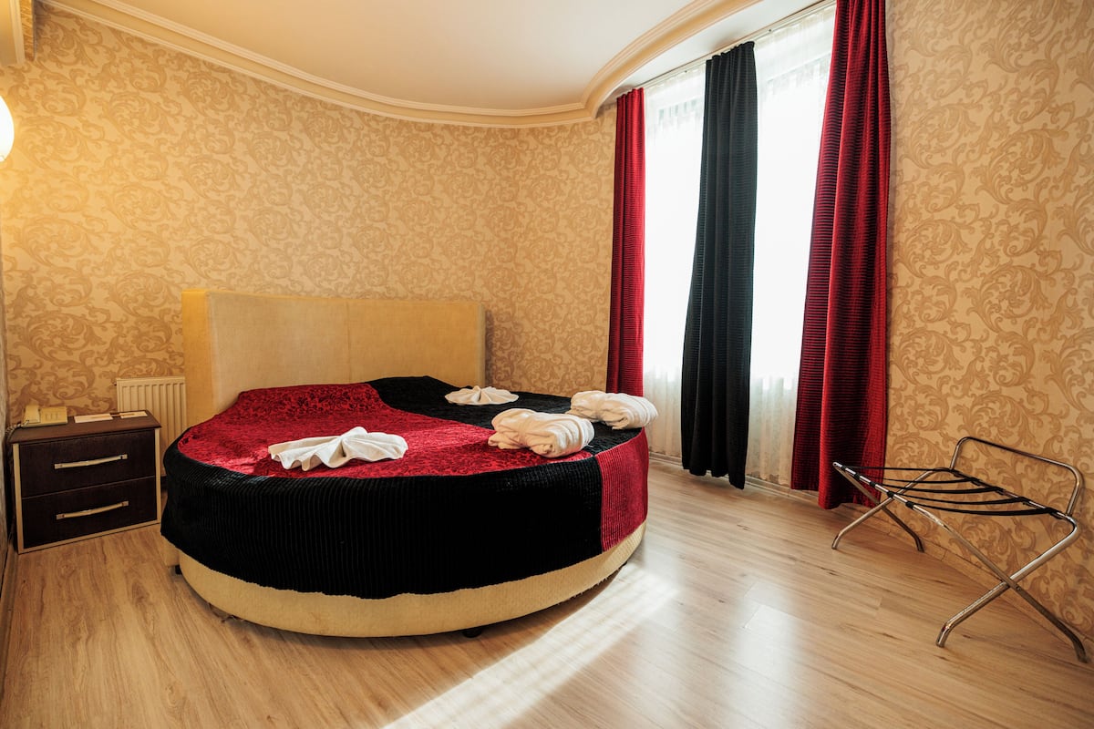 Ein Hotelzimmer mit einem runden Bett, einem Nachttisch, einem Heizkörper und einem Fenster mit roten Vorhängen.