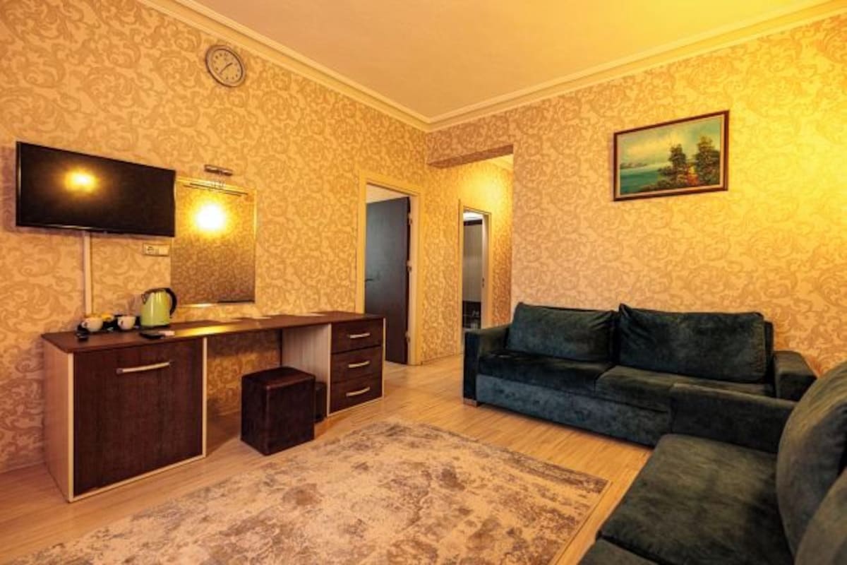 Un salon avec un canapé vert foncé, un téléviseur à écran plat fixé au mur, un meuble en bois avec des tiroirs et un tapis à motifs sur le sol.