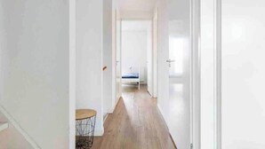 A narrow hallway with a wooden floor leading to a bedroom with a bed and a small round table.