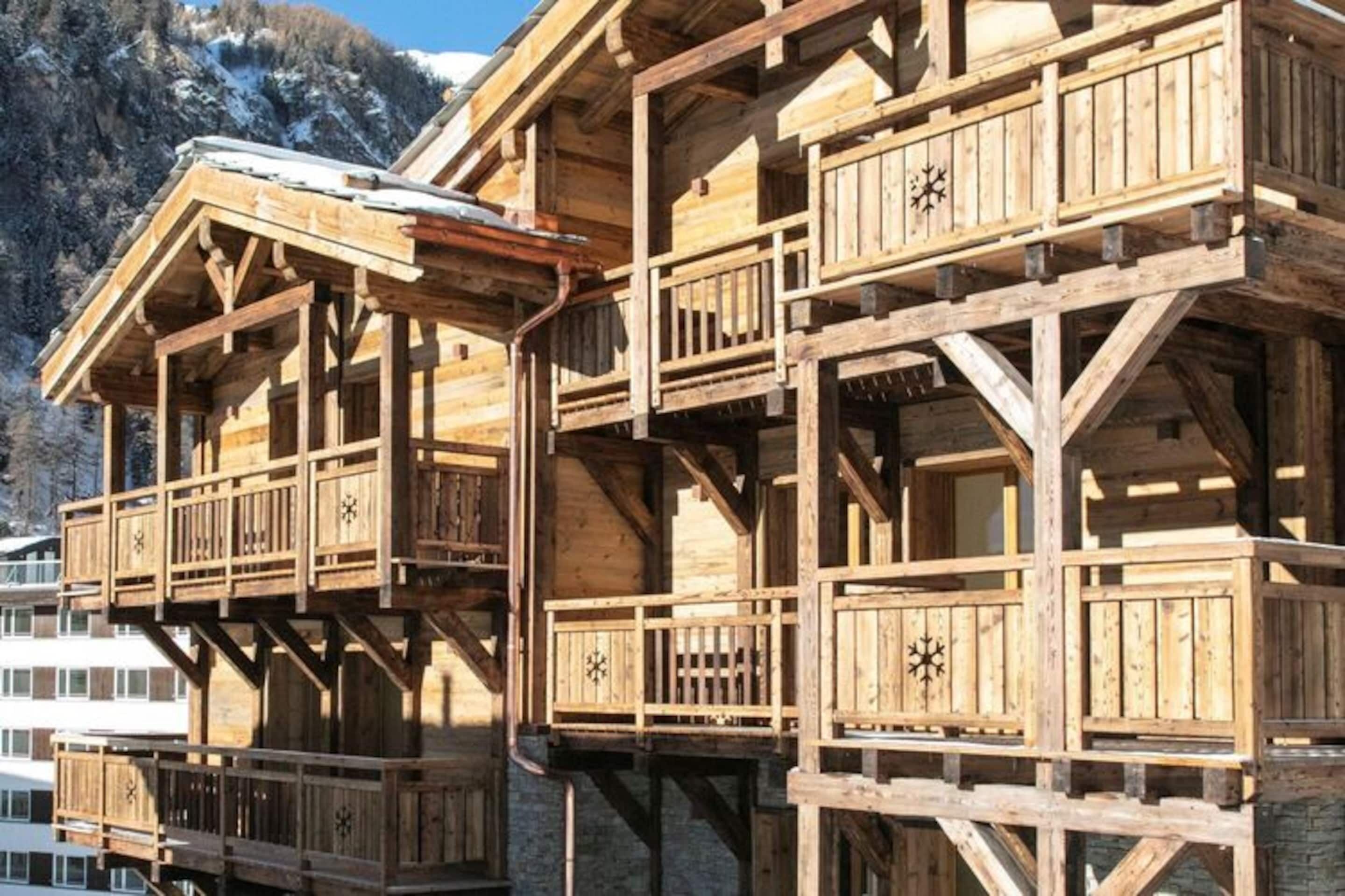 A wooden chalet with multiple balconies and a stone foundation, set against a mountainous backdrop.