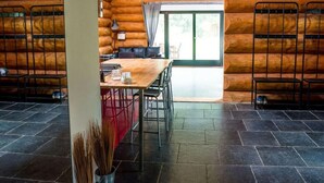 Interior de una cabaña de troncos con una mesa de comedor de madera, sillas metálicas, un sofá y un ventanal grande.