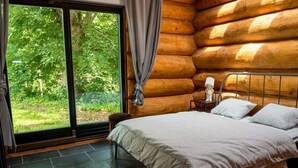 Un dormitorio de cabaña de troncos con cama, mesita de noche y vistas al follaje a través de un ventanal grande.