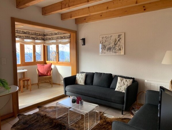 A cozy living room with a dark grey sofa, a clear coffee table, a wooden chair, and a view of the outdoors through large windows.