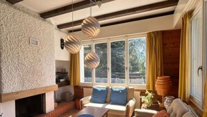A cozy living room with a fireplace, a sofa, a dining table, and a large window with curtains.