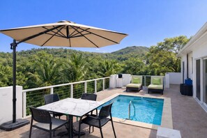 A rooftop pool with a table and chairs, a lounge area with cushions, and a view of a forested hill.