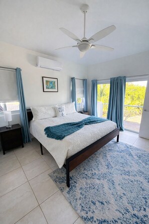 A bedroom with a bed, a ceiling fan, a nightstand, a window with curtains, and a view of greenery outside.