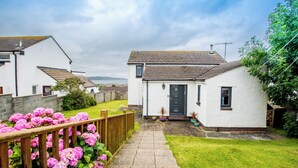 A white house with a grey roof, a front door, and a small garden with pink flowers. - PuffinPatch - Beachside Cottage (Broad Haven)