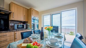 A modern kitchen with wooden cabinets, a built-in oven, and a dining area with a table set for two. - PuffinPatch - Beachside Cottage (Broad Haven)