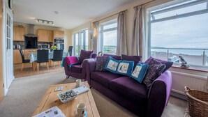 A modern living room with a purple sofa, a dining area, and a kitchen. - PuffinPatch - Beachside Cottage (Broad Haven)