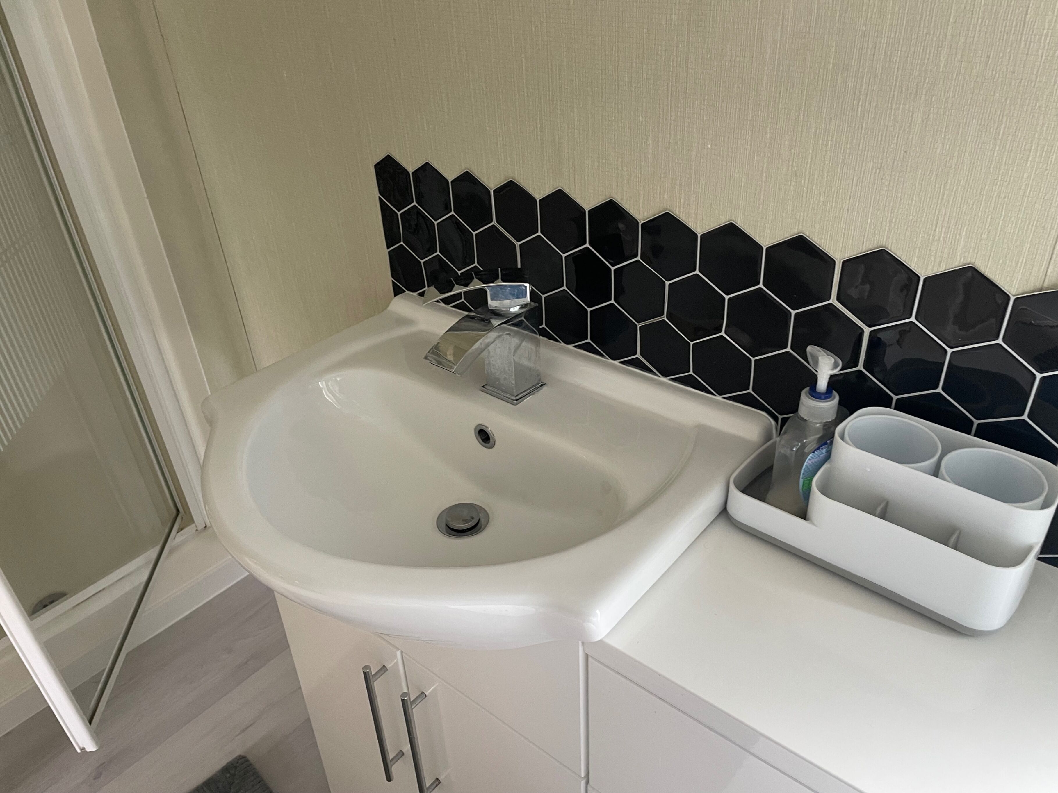 A bathroom with a white sink, a black hexagonal tile backsplash, and a cup holder with a bottle.