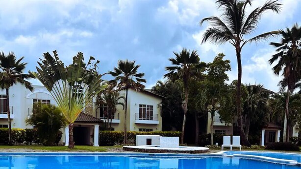 A swimming pool with lounge chairs facing the water, surrounded by palm trees and a building with balconies. - Carey Apartment in Carey Residential, White Sands, 5 Minutes from the Beach. (Punta Cana)