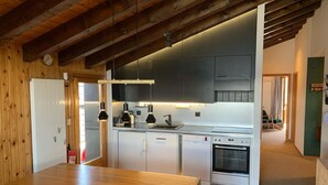 A modern kitchen with wooden beams, a dining area with a wooden table and red chairs, and a view into a living area.