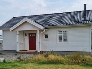 Ett envåningshus med en röd dörr, vit träpanel och ett grått tak täckt med takpannor.