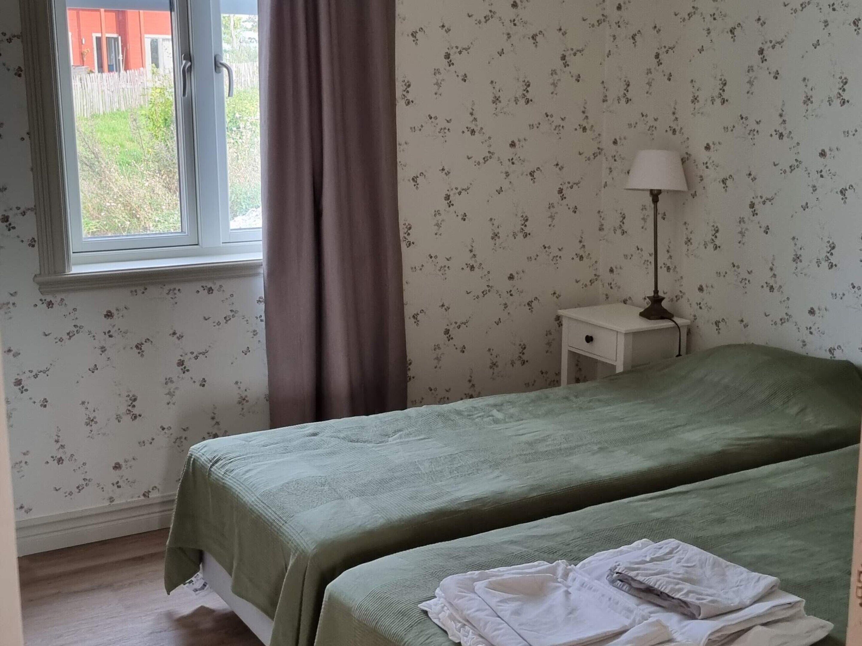 A room with two beds, a window with curtains, a nightstand with a lamp, and floral wallpaper.