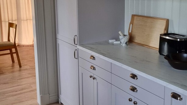 Une cuisine avec des meubles blancs, un plan de travail en marbre, une chaise en bois et un grille-pain.