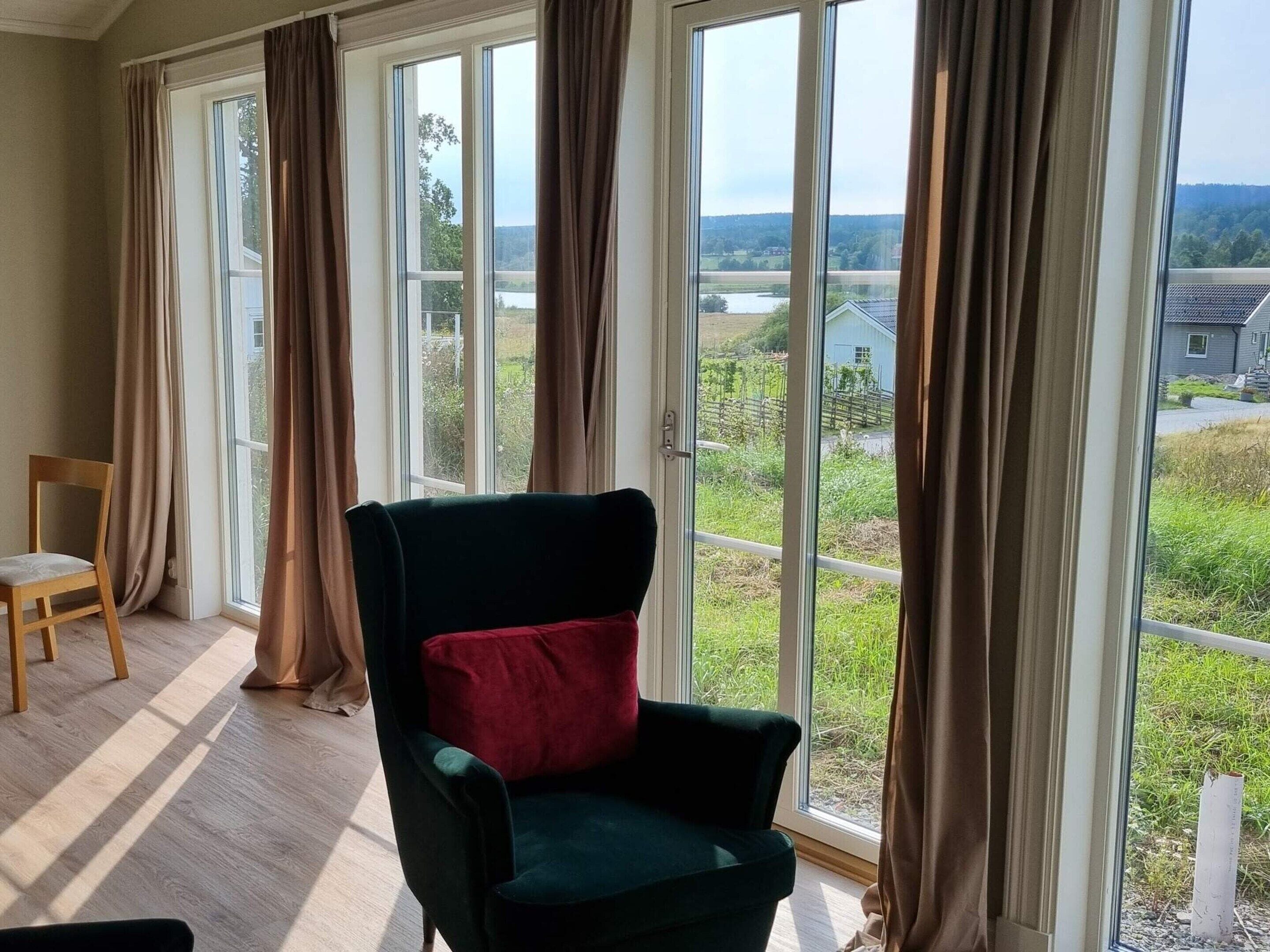 Una habitación con un ventanal grande, una silla verde con un cojín rojo y otra silla de madera.