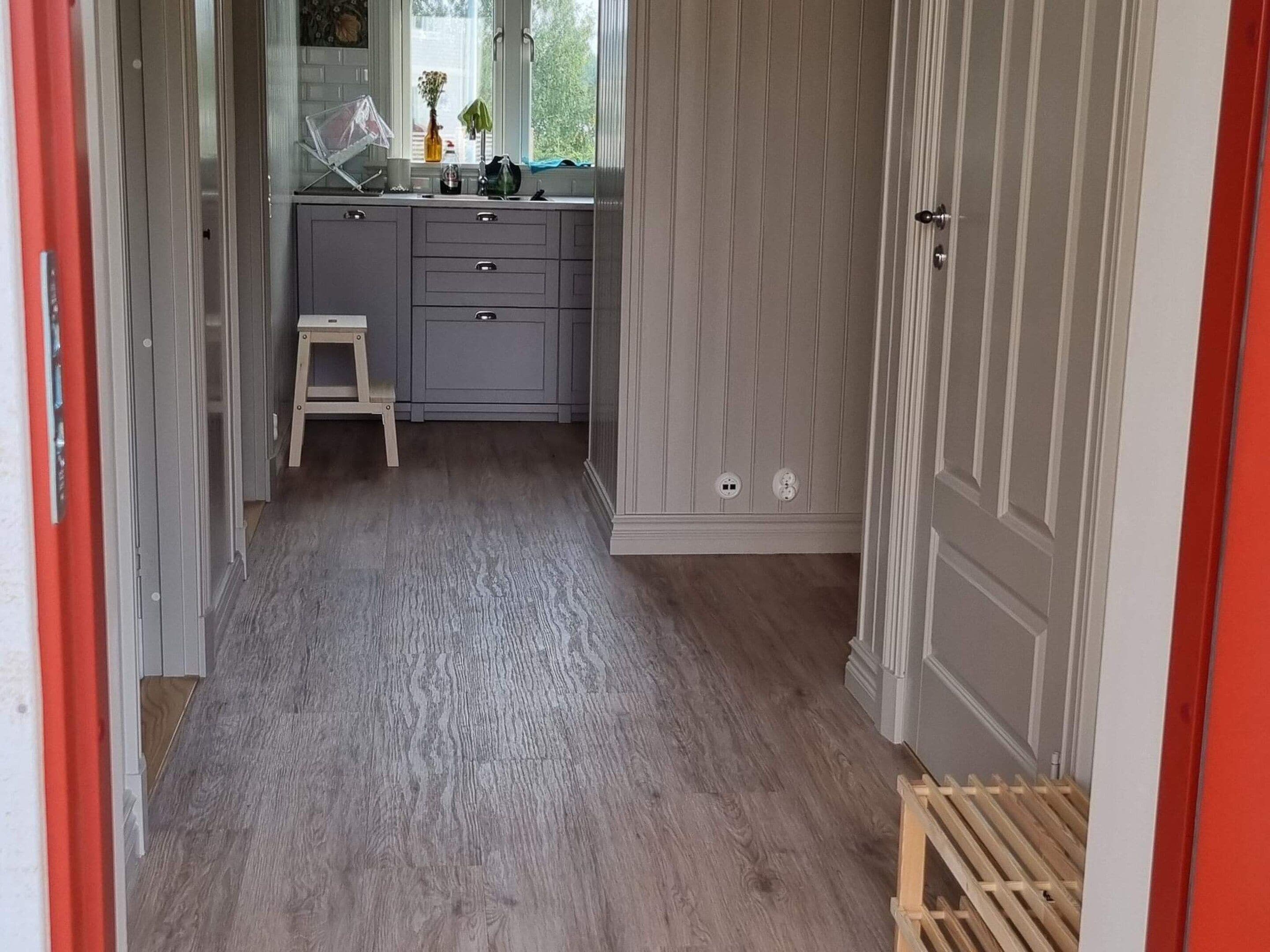 A narrow hallway with a grey wooden floor leading to a kitchen area with grey cabinets and a small stool.