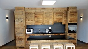 A modern kitchen with wooden cabinetry, a black countertop, a microwave, a coffee maker, and three white stools. - Spacious Apartment for ski Holiday (Veysonnaz)